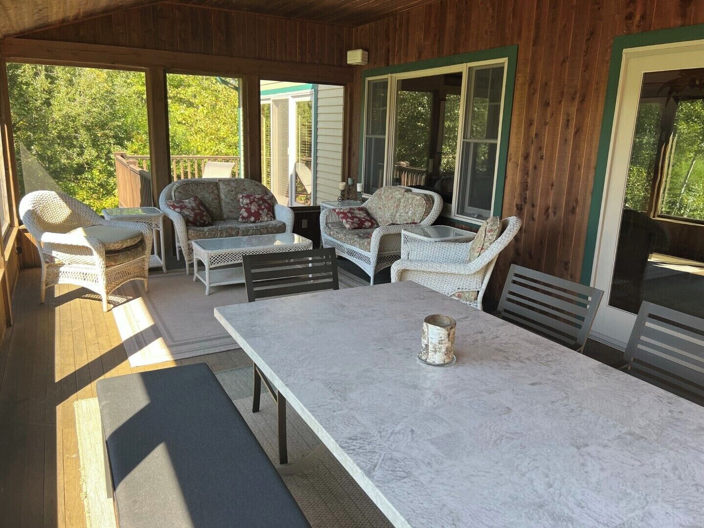 Eine überdachte Terrasse mit Korbmöbeln, einem Marmortisch und großen Fenstern.