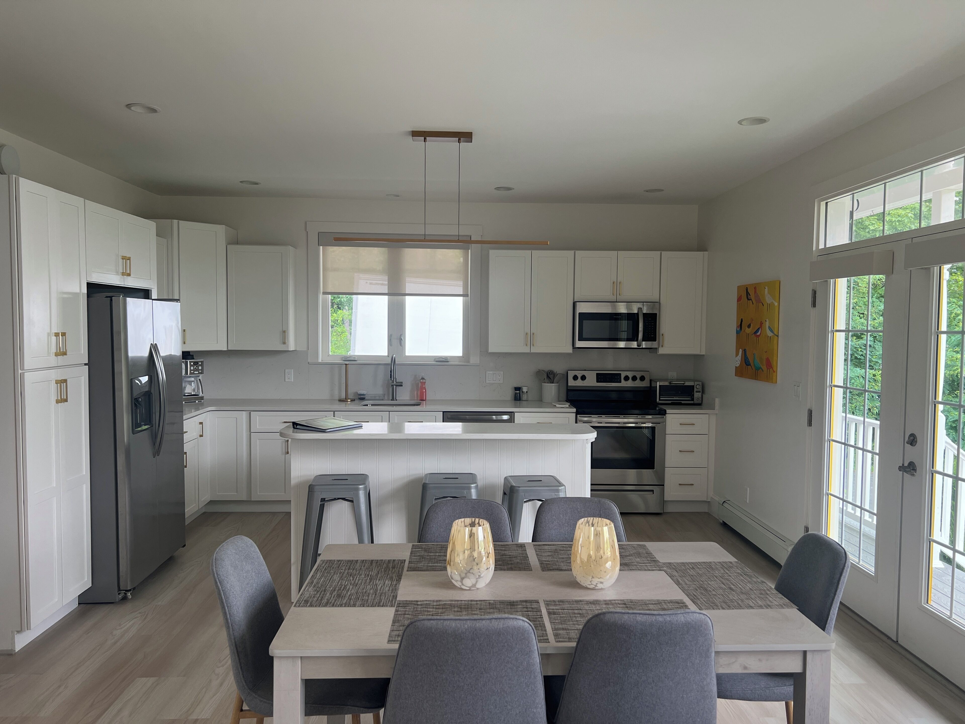 A modern kitchen with white cabinetry, stainless steel appliances, a central island, and a dining area with gray chairs and a table.