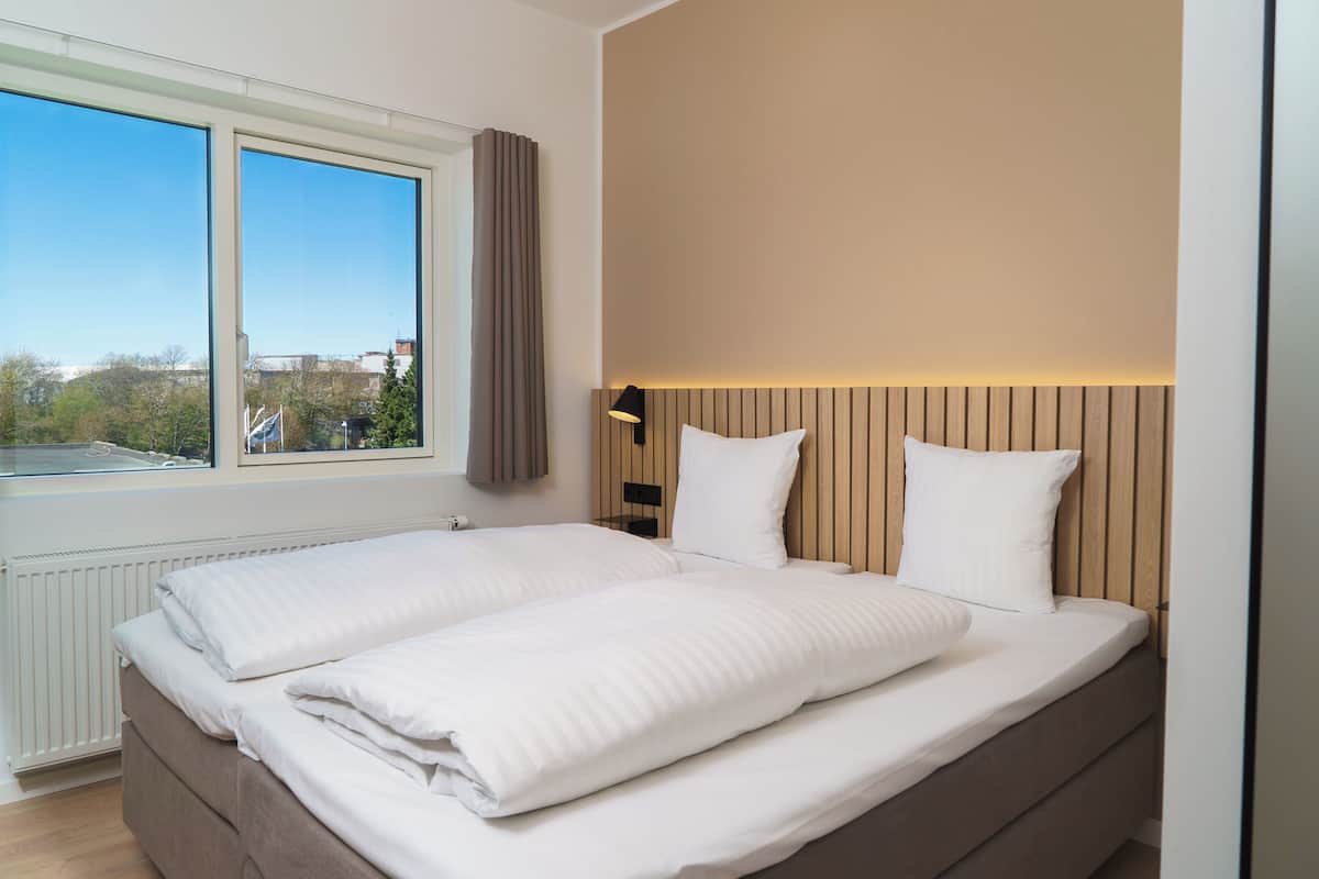 A double bed with white linens, a wooden headboard, a bedside table with a lamp, and a window with curtains.