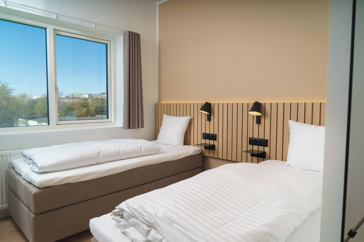 A hotel room with two beds, a window with curtains, and wall-mounted lamps.