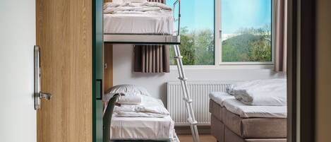 A room with a bunk bed, a single bed, a desk, and a window with a view of greenery.