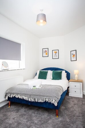A bedroom with a bed, a bedside table, a lamp, a radiator, and framed artwork on the wall.