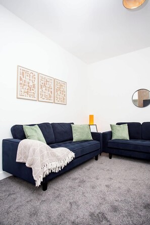 A modern living room with two dark blue sofas, a patterned throw blanket, and a grey carpet.