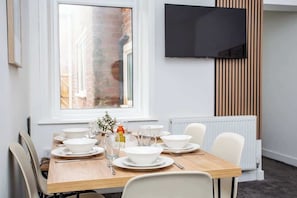 A dining area with a wooden table set for four, white chairs, a TV mounted on the wall, and a window with a view of a brick building.
