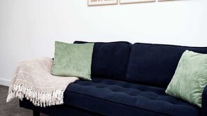 Ein dunkelblaues Sofa mit einer weißen, gemusterten Decke und zwei Kissen, eines grün und eines weiß, vor einer schlichten Wand mit drei gerahmten abstrakten Kunstwerken.
