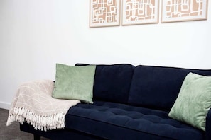 A dark blue sofa with a white patterned throw blanket and two pillows, one green and one white, against a plain wall with three framed abstract art pieces.