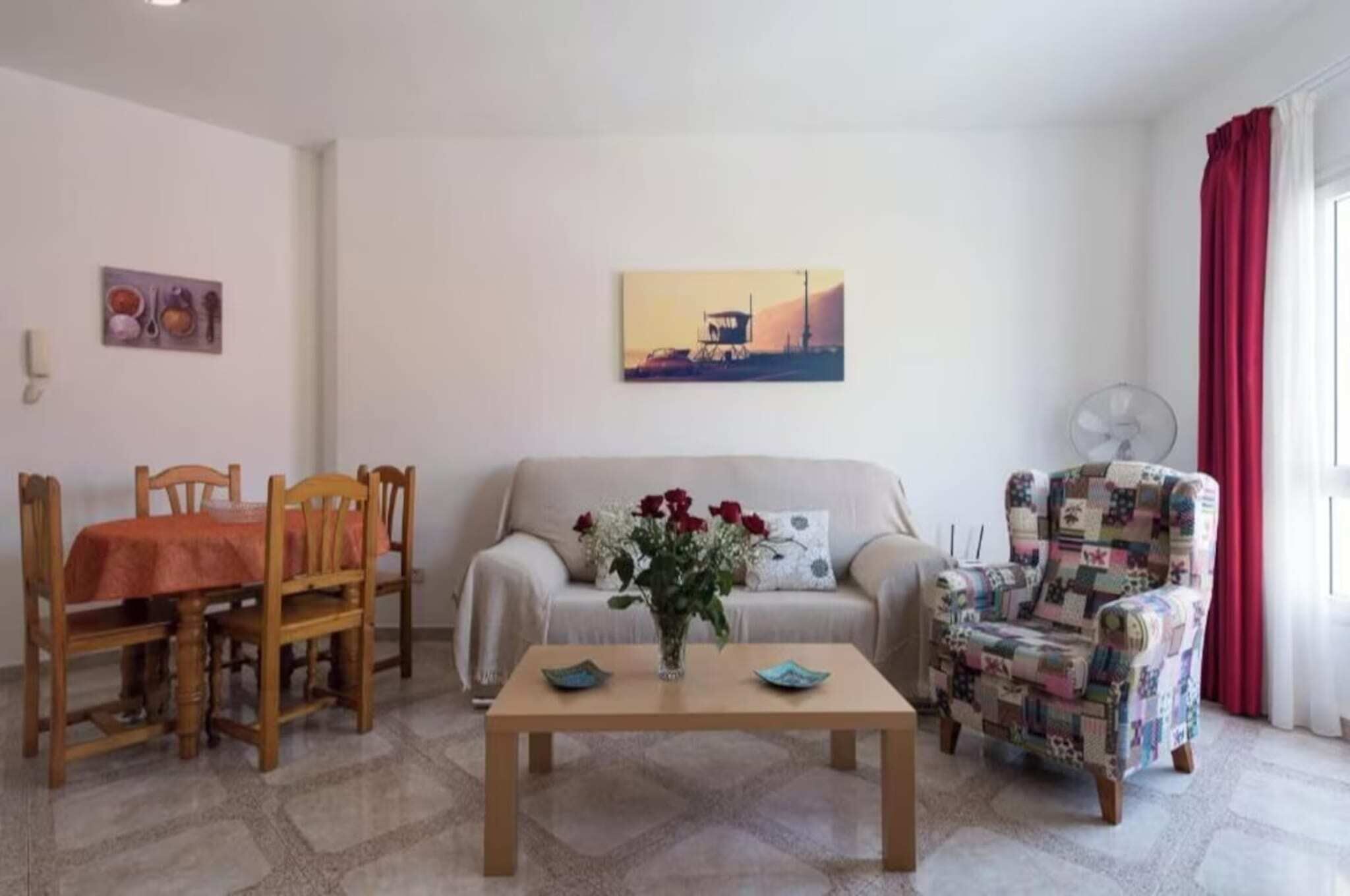 Un salon comprenant un canapé, une table basse, une table à manger avec des chaises, un fauteuil à motifs, un tableau accroché au mur et une fenêtre avec des rideaux.