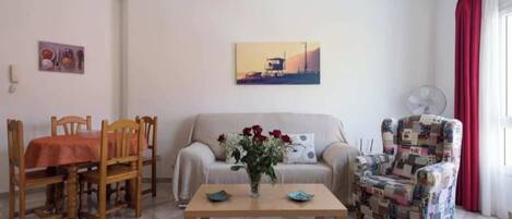 Un salon comprenant un canapé, une table basse, une table à manger avec des chaises, un fauteuil à motifs, un tableau accroché au mur et une fenêtre avec des rideaux.