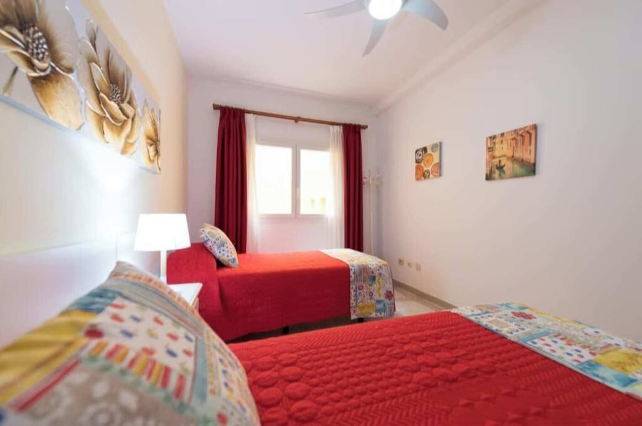 A room with two beds, a window with red curtains, a ceiling fan, and wall art.