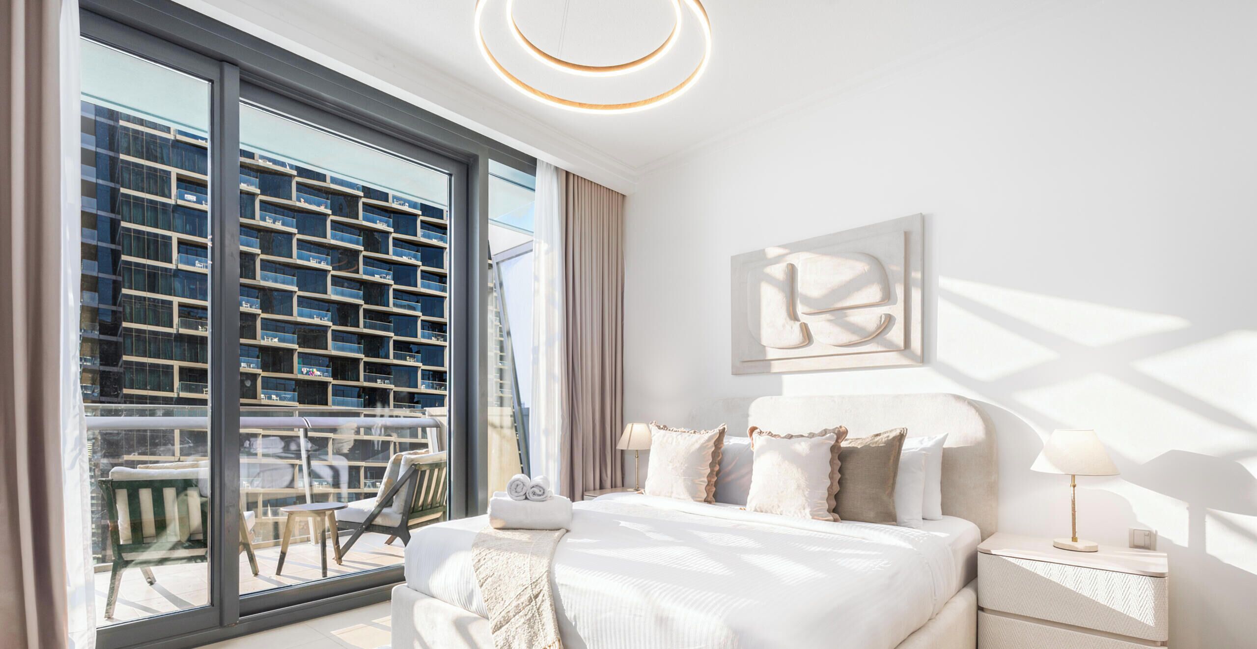 A modern hotel room with a large window, a bed with pillows, a bedside table with a lamp, and a wall-mounted abstract art piece.