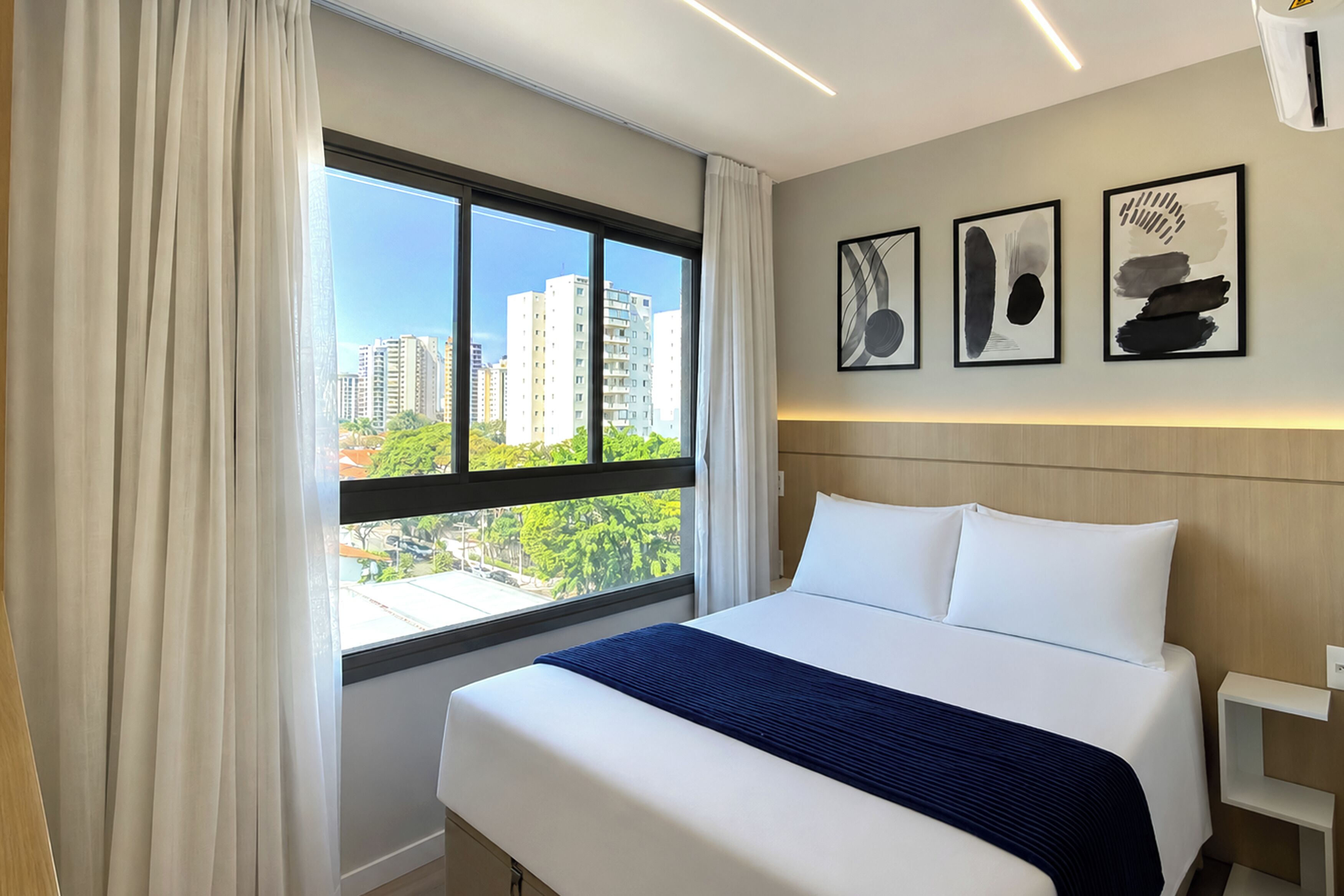 Ein Hotelzimmer mit einem großen Bett, Blick auf die Stadt durch ein Fenster und drei abstrakte Kunstwerke an der Wand.