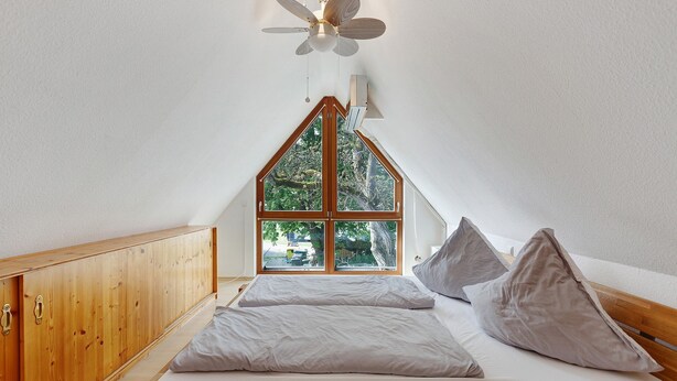 Un dormitorio con ventilador de techo inclinado, armario de madera, cama con almohadas y ventana con vistas a la vegetación.