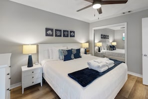 A bedroom with a bed, two bedside tables, a ceiling fan, wall art, and a mirror. - New Listing! Cozy 2 bed 1.5 bath at Gulf Highlands (Panama City Beach)