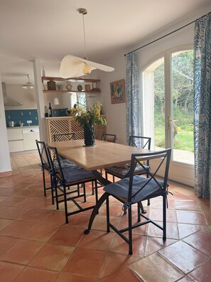 A dining area with a wooden table and chairs, a kitchen in the background, and a view of the outdoors through large windows.