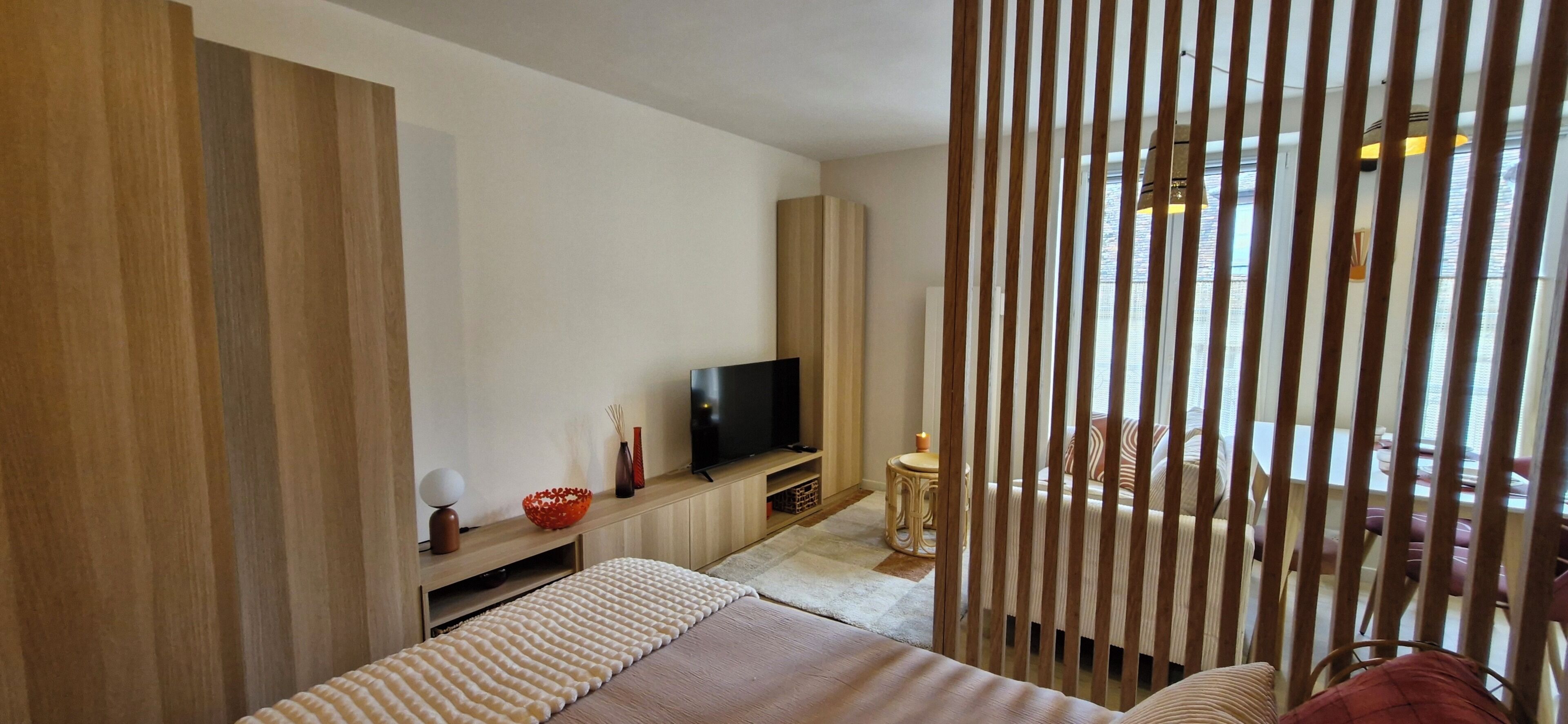 A modern hotel room with a TV, wooden paneling, a dining area, and a view through a window.