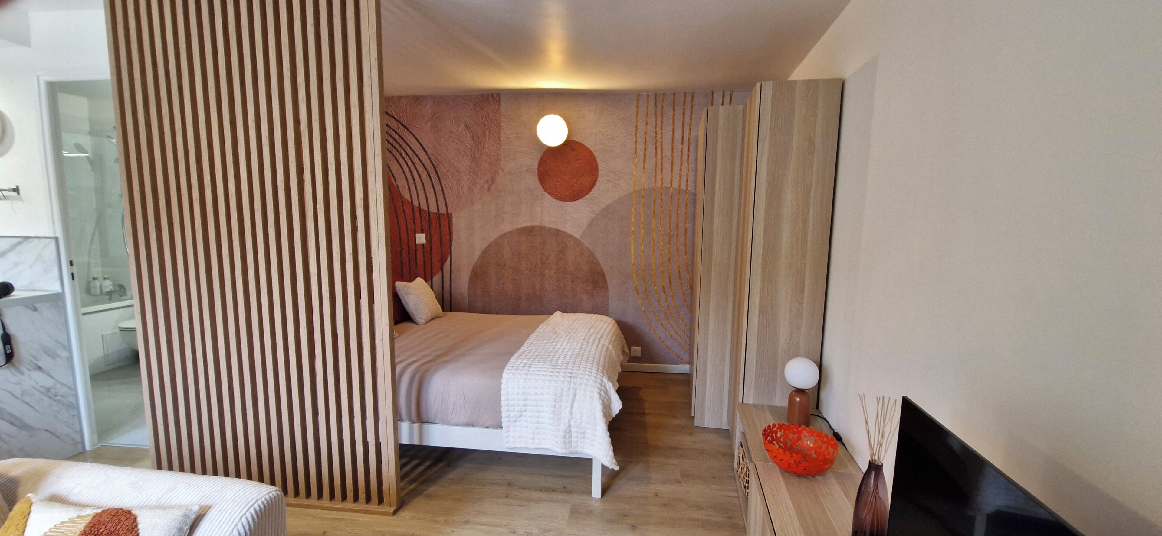 Un dormitorio moderno con una cama, un panel de madera, una pared con arte abstracto, una lámpara redonda y un televisor sobre un pedestal de madera.