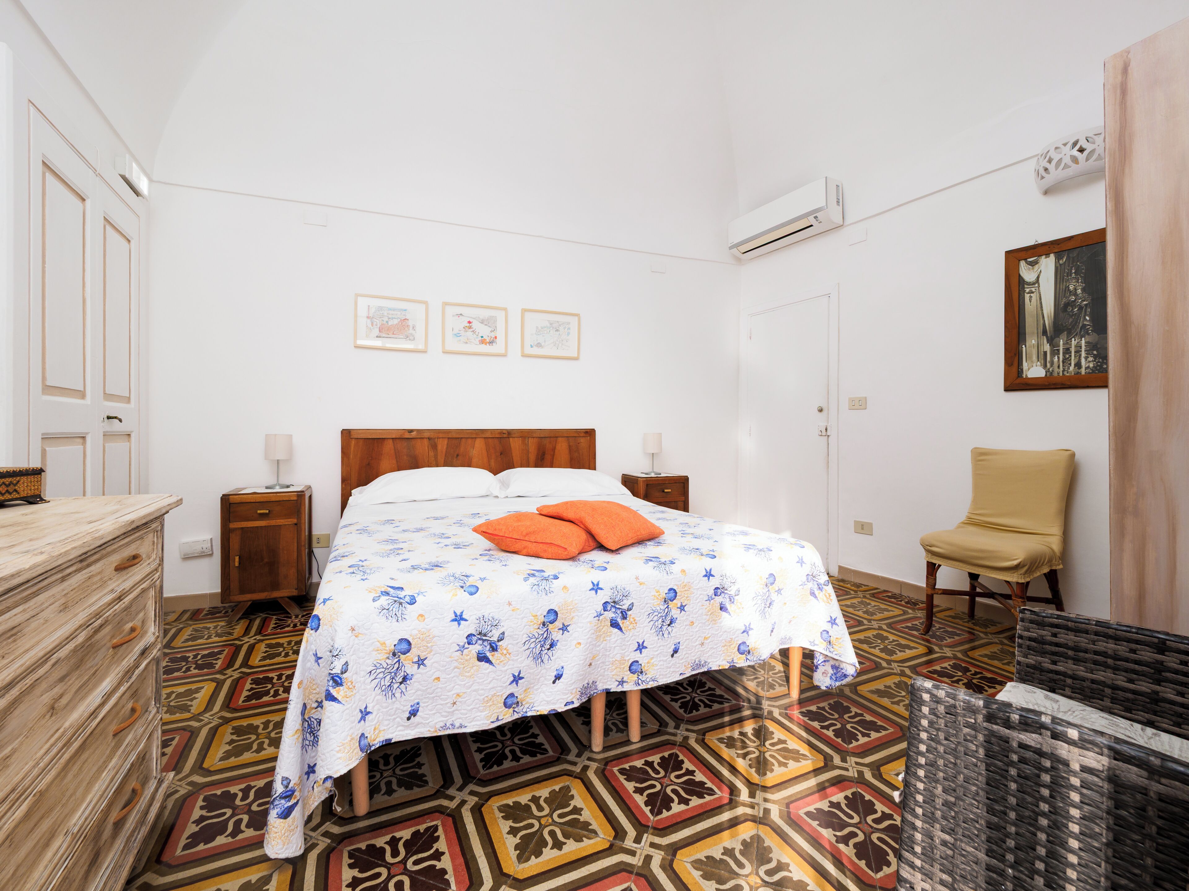 Quarto com cama, duas mesas de cabeceira, uma cadeira, cômoda e piso estampado.