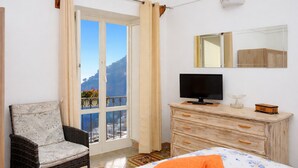 A bedroom with a bed, a chair, a dresser, a TV, and a window with a view of a mountain. - De Vivo Realty - La casa del Cavaliere (Positano)