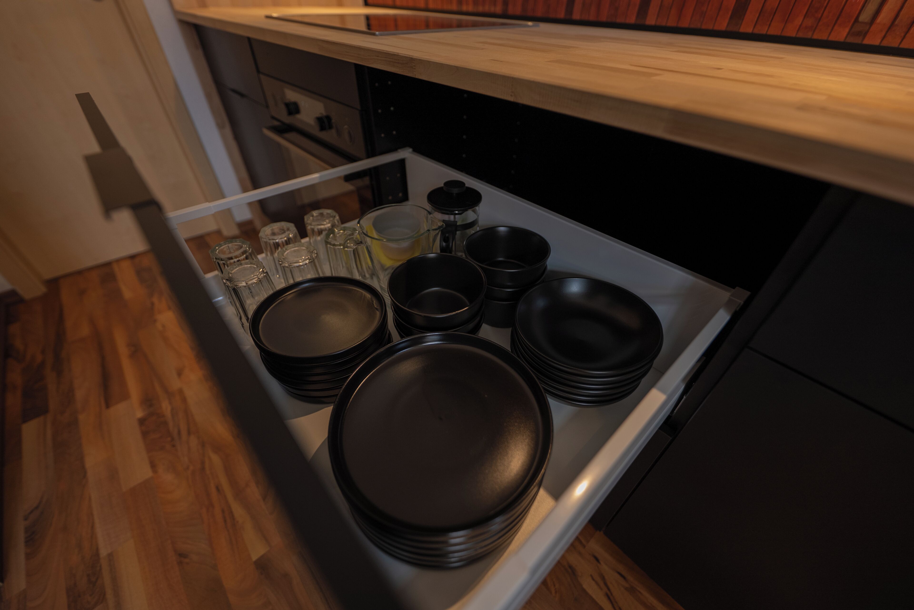 A kitchen with a drawer containing black plates, glasses, and a bottle.
