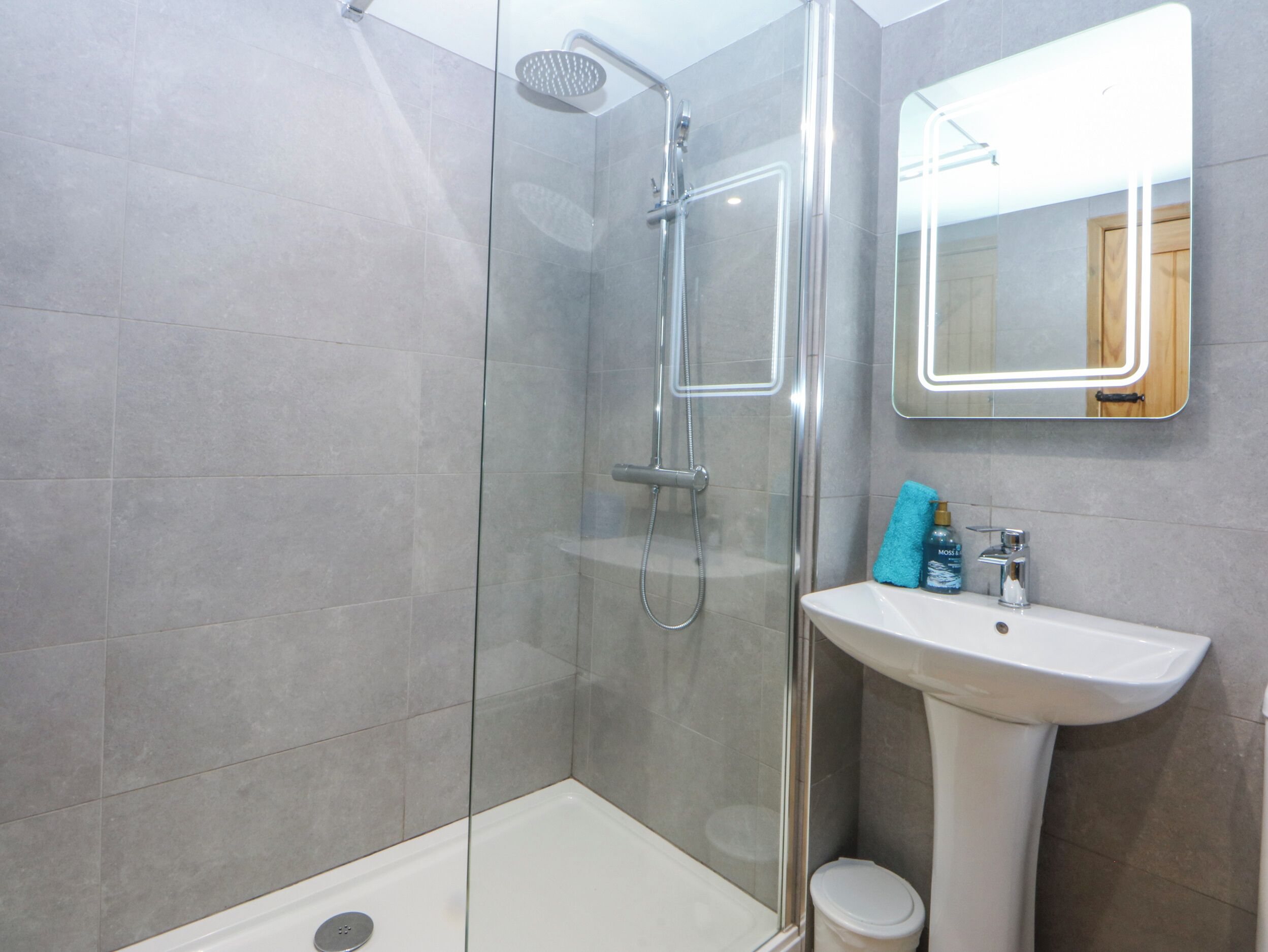 Un baño moderno con ducha envuelta en vidrio, un lavabo blanco y un espejo con marco iluminado.