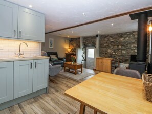 A kitchen with light blue cabinets, a wooden dining table, a stone wall, a fireplace, and a cozy seating area with a sofa and armchair. - Nyth Dryw (Criccieth)