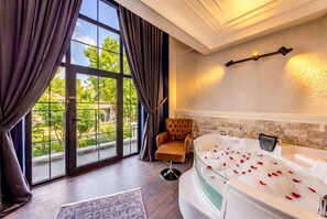 Una habitación con un ventanal grande, un sillón de cuero, una bañera con pétalos de rosa y una lámpara fijada a la pared.