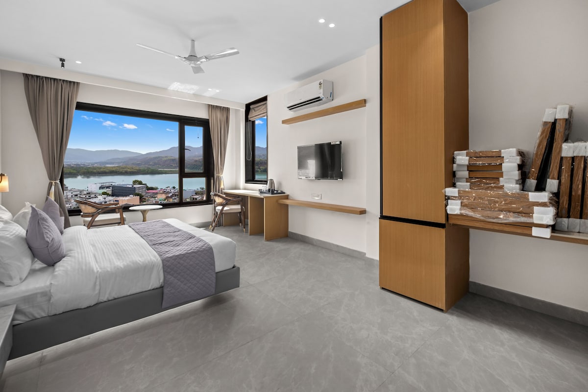 Ein modernes Hotelzimmer mit Bett, Schreibtisch, Sessel, Fernseher und einem großen Fenster mit Blick auf einen See und Berge.