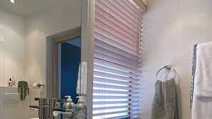 Un baño con un lavamanos grande de color blanco, un espejo, un perchero para toallas y una ventana con persianas.