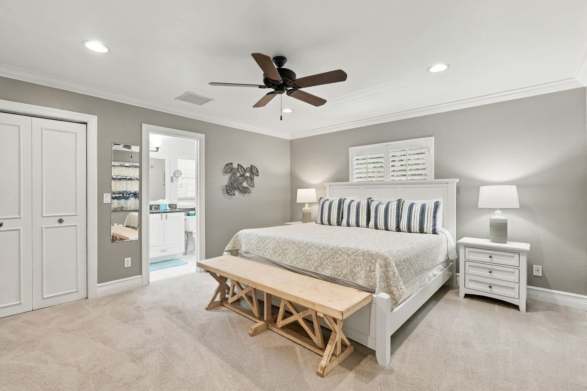 A bedroom with a bed, bedside tables, a bench, a ceiling fan, and a door leading to a bathroom.