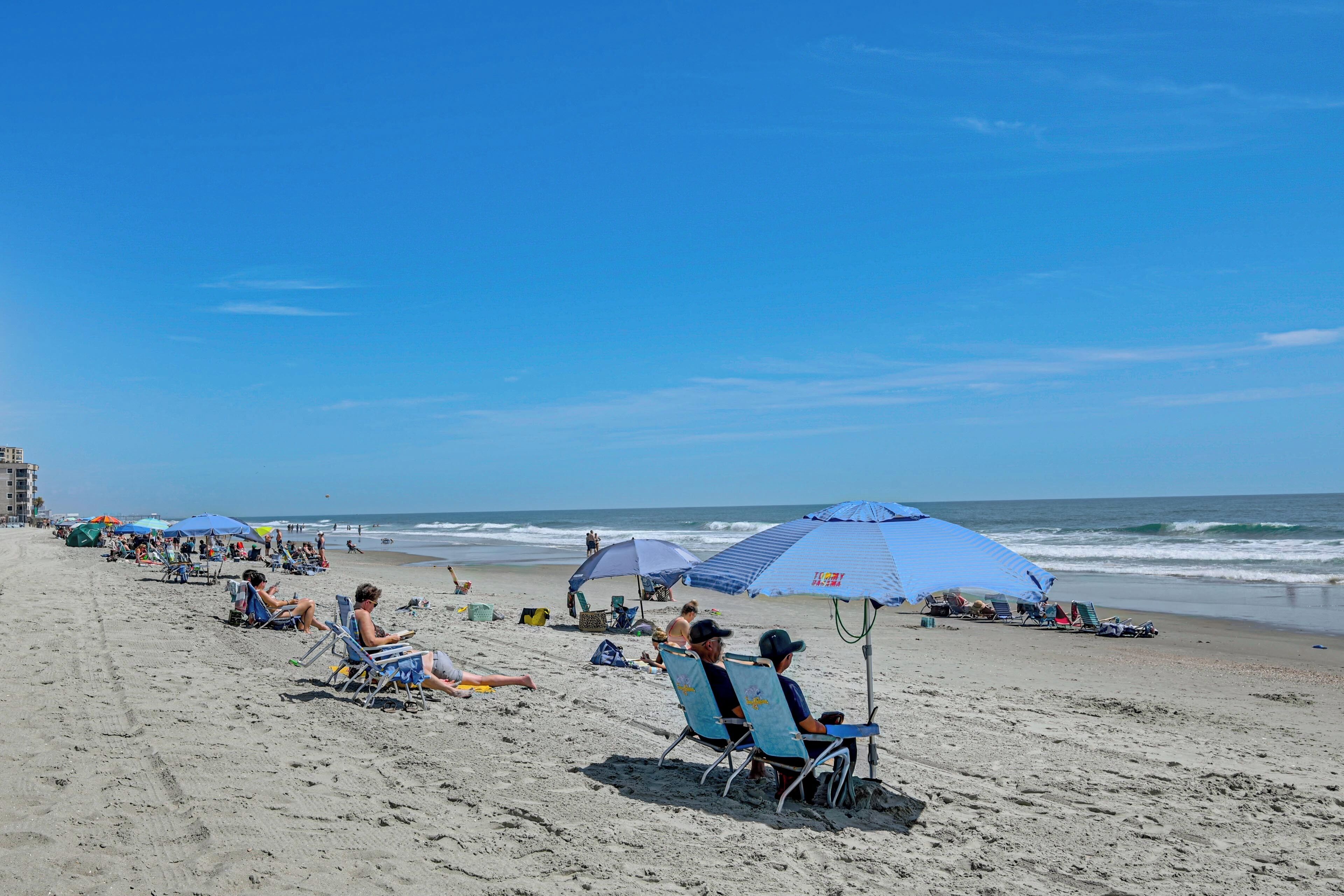 Una playa con gente relajándose bajo sombrillas, algunos en sillas y otros de pie.