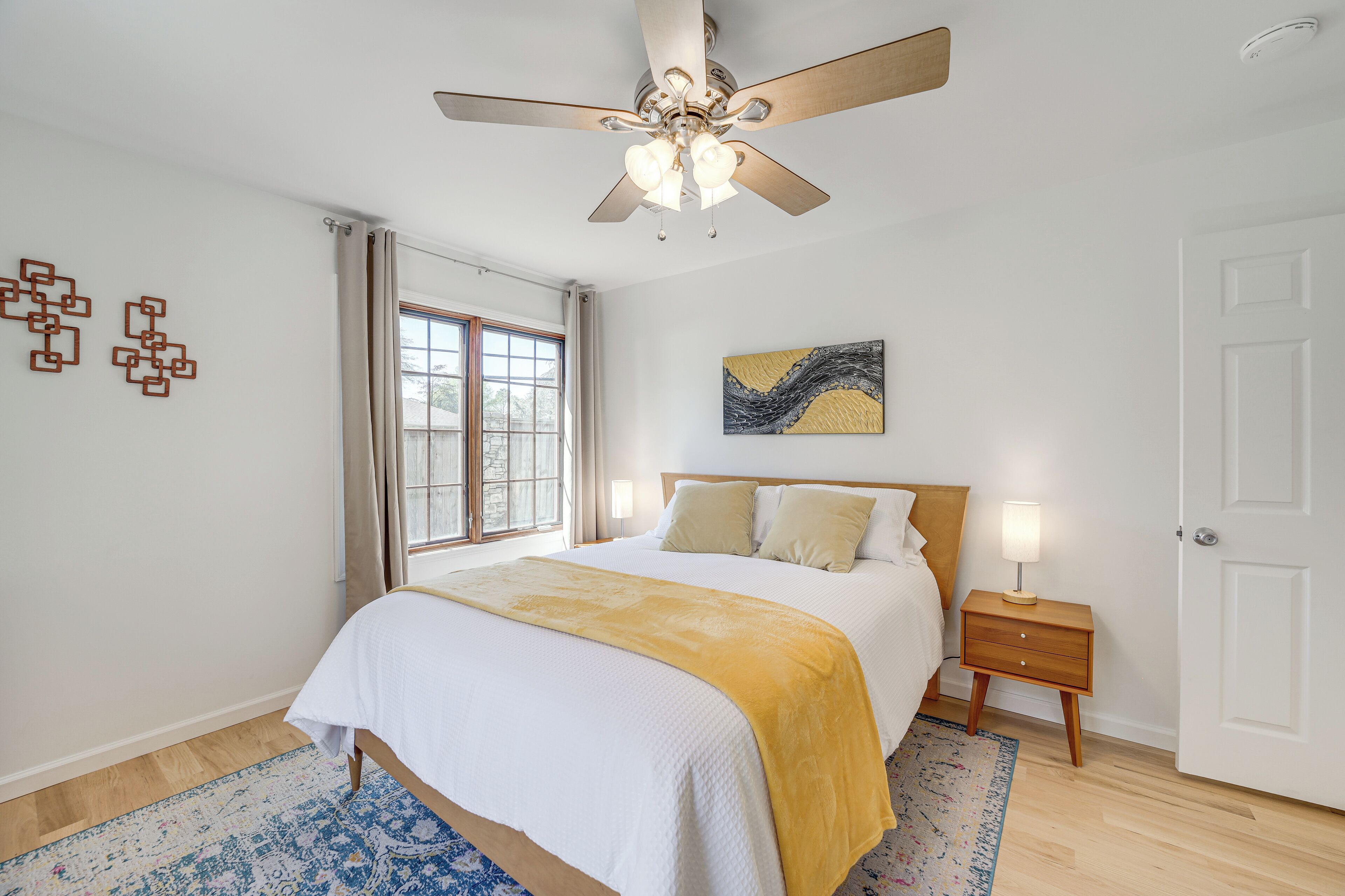 A bedroom with a bed, a ceiling fan, a nightstand, a lamp, a window with curtains, and a door.