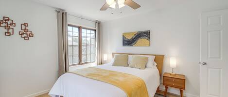A bedroom with a bed, a ceiling fan, a nightstand, a lamp, a window with curtains, and a door.