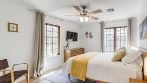 Un dormitorio con cama, una silla, ventilador de techo, televisor y ventana.