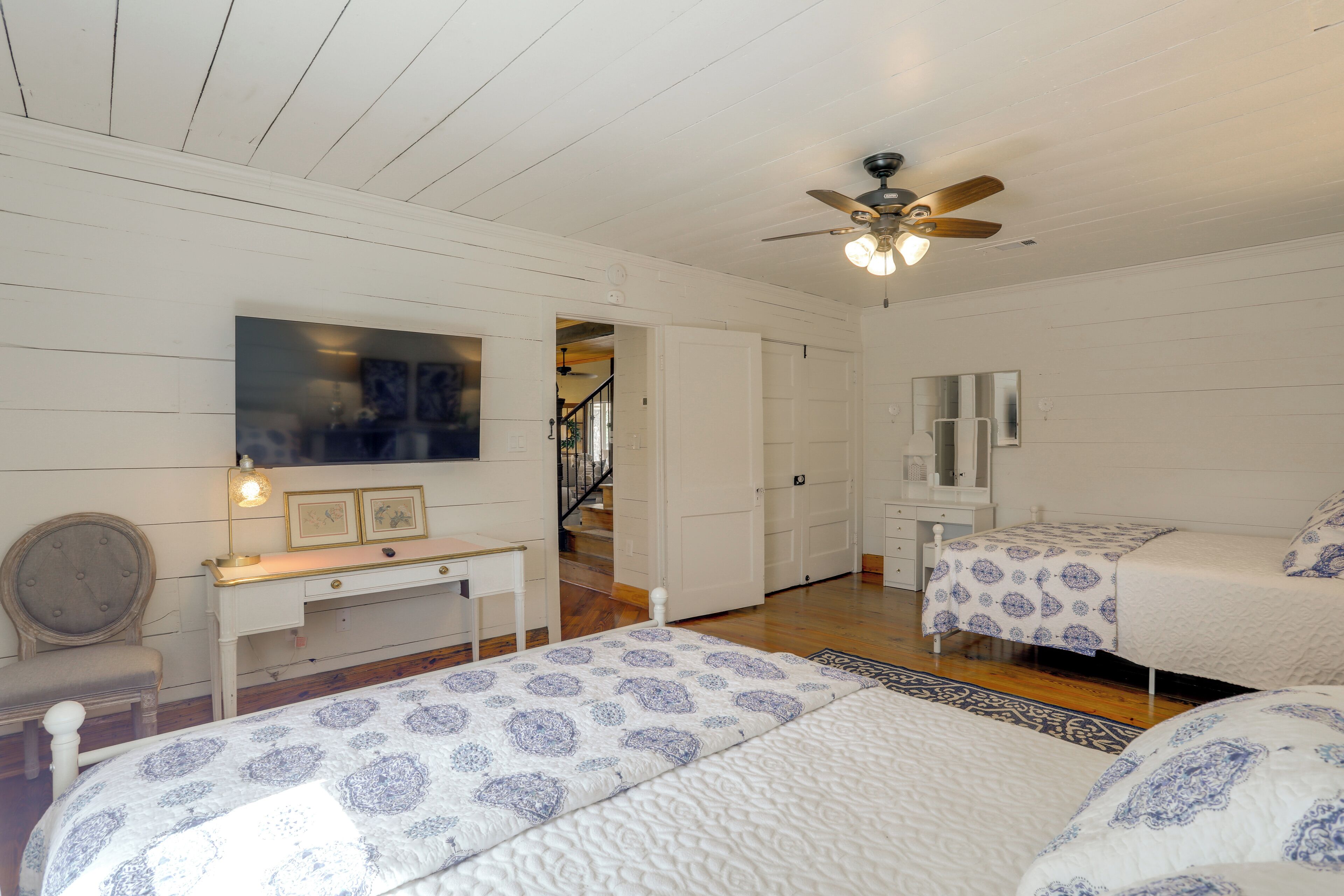 A bedroom with two beds, a desk, a chair, a ceiling fan, and a staircase.
