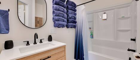 A bathroom with a sink, mirror, towels, shower curtain, and a bench.
