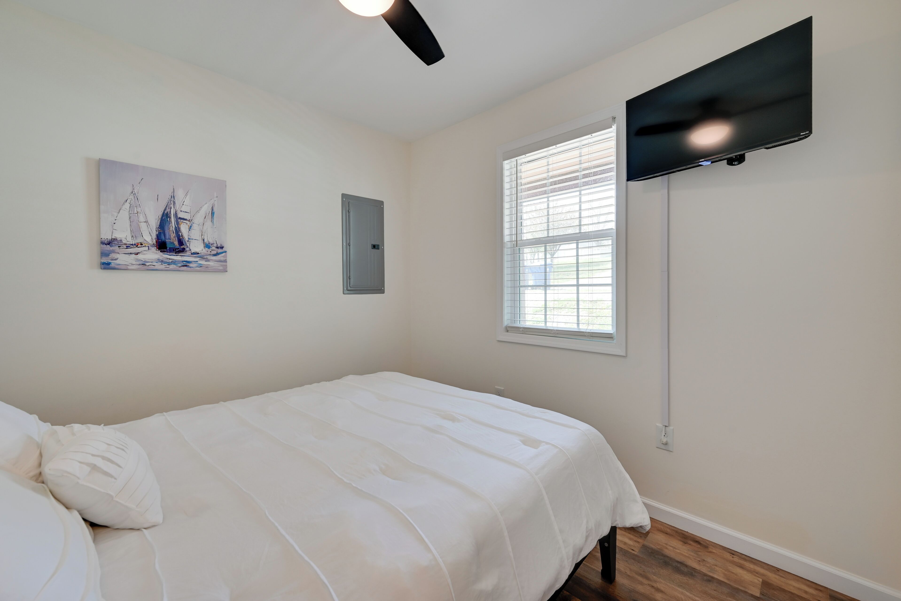 A bedroom with a bed, a ceiling fan, a TV mounted on the wall, a window with blinds, and a painting on the wall.