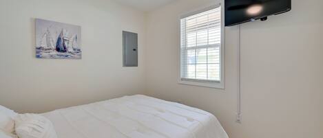 A bedroom with a bed, a ceiling fan, a TV mounted on the wall, a window with blinds, and a painting on the wall.