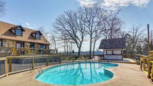 Piscina com design curvo, área de deck cercada e um edifício com telhado inclinado ao fundo.