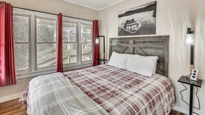 Ein Schlafzimmer mit großem Fenster, einem Bett mit kariertem Überwurf, einem Holz-Kopfteil, einem Nachttisch mit Lampe und einem Wandbild.
