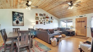 A spacious living room with a wooden ceiling, a dining table with chairs, a sofa, a fireplace, and a wall adorned with various framed pictures. - Walk to Private Lake! Family Cabin w/ Large Decks (Show Low)