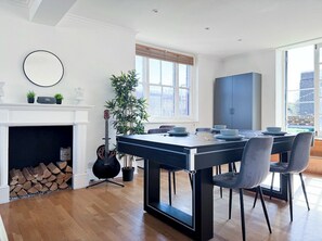 A modern dining area with a large table, chairs, a fireplace, a guitar, and a potted plant. - Spacious 2-Bedroom Flat in Fulham | 85” TV, PS5, Pool Table & Terrace (London)