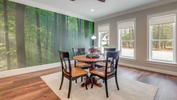 A dining area with a round table, four chairs, a ceiling fan, and a large window with a forest mural.