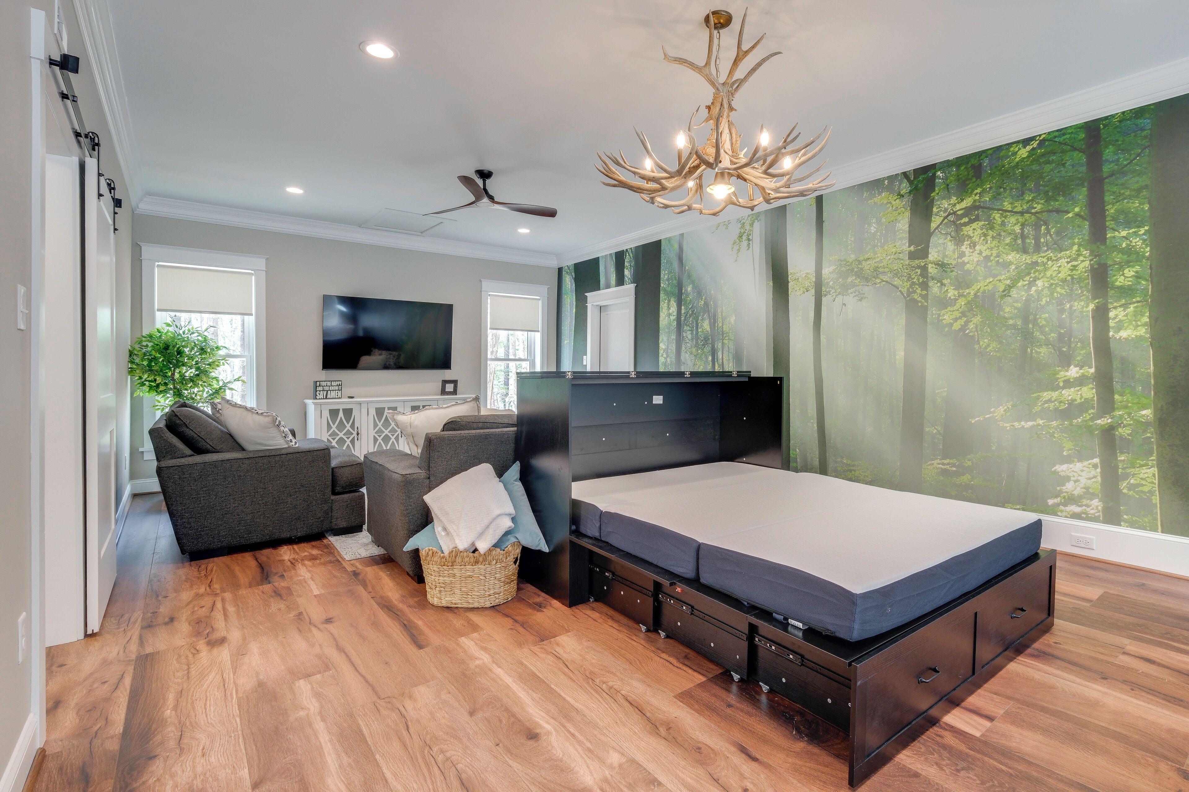 A modern bedroom with a bed, a ceiling fan, a chandelier, and a large forest mural.