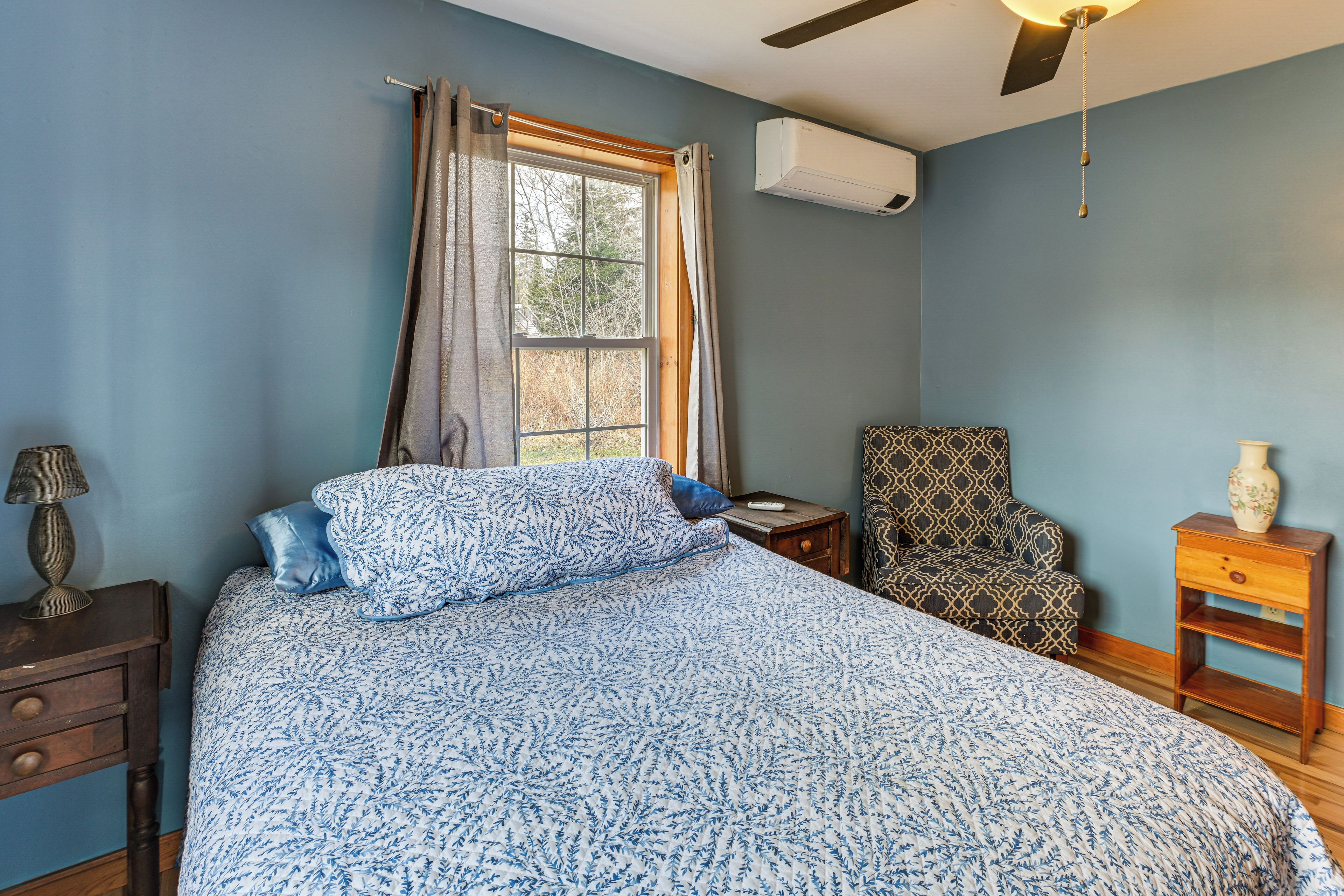 Une chambre à coucher comprenant un lit, une chaise, une table de chevet, une lampe, une fenêtre avec des rideaux et un appareil de climatisation.
