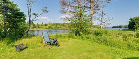 A grassy area with a bench and a chair, a body of water, and trees in the background.