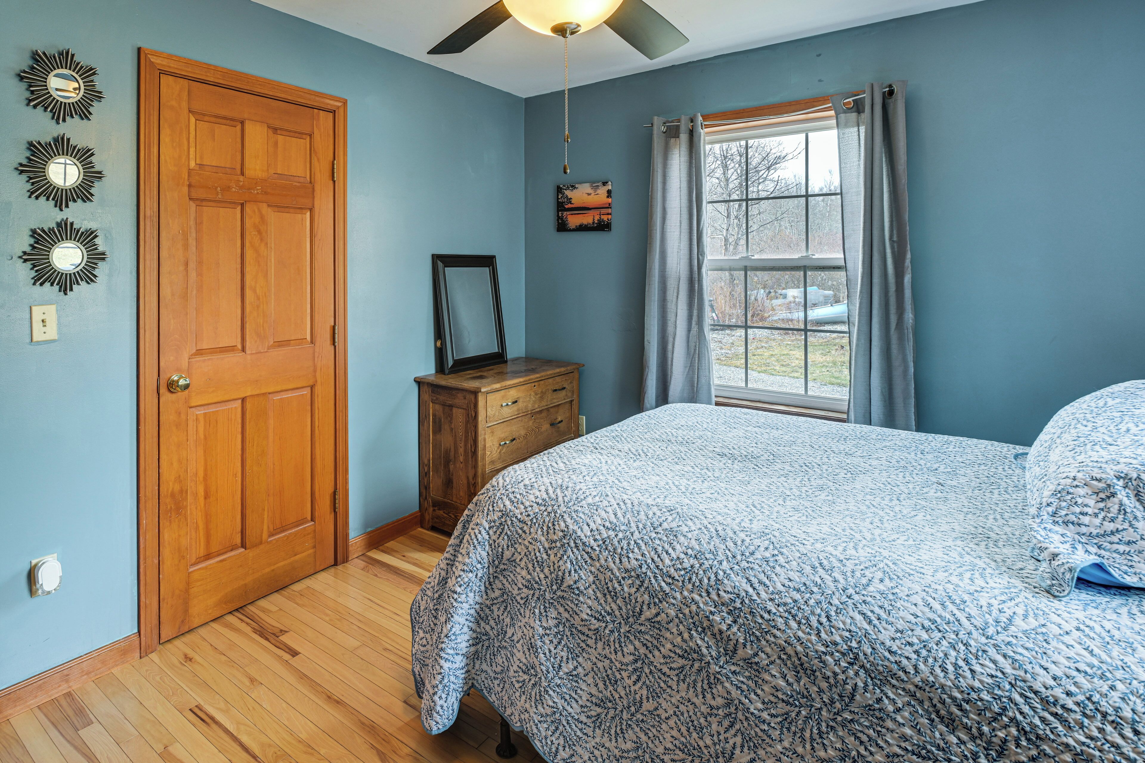 A bedroom with a wooden door, a bed with a patterned bedspread, a dresser, a ceiling fan, and a window with curtains.