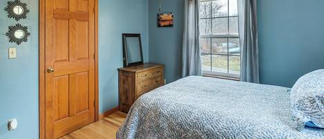 A bedroom with a wooden door, a bed with a patterned bedspread, a dresser, a ceiling fan, and a window with curtains.