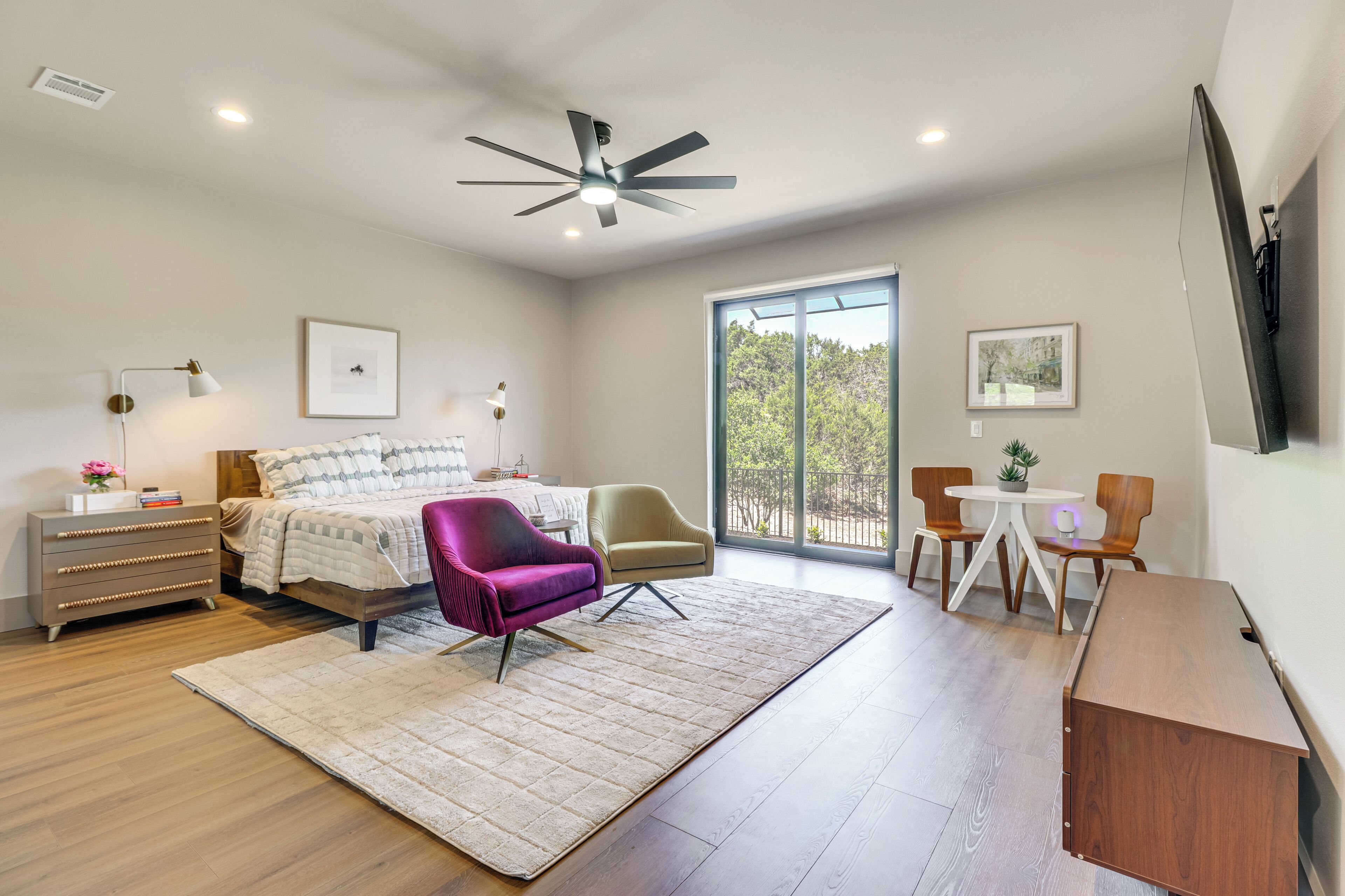 A modern bedroom with a bed, a desk, chairs, a ceiling fan, and a large window.