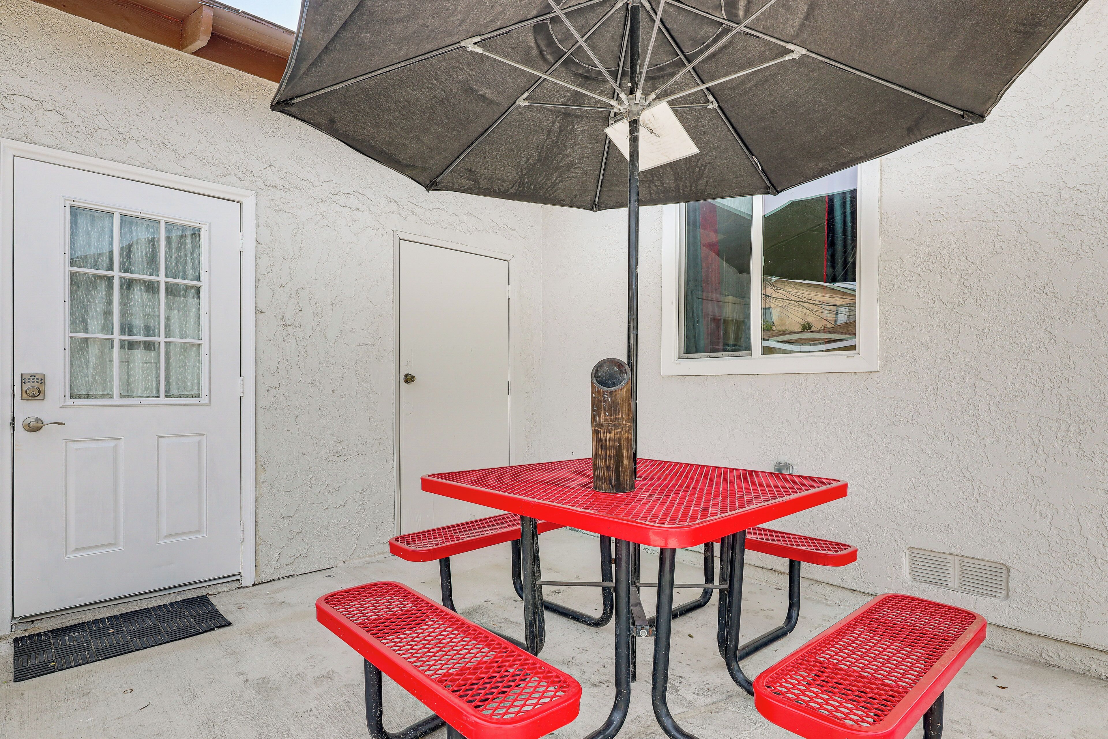 En terrasse med et rødt metalbord og bænke, en parasol, en hvid dør og et vindue med udsigt til en bygning.
