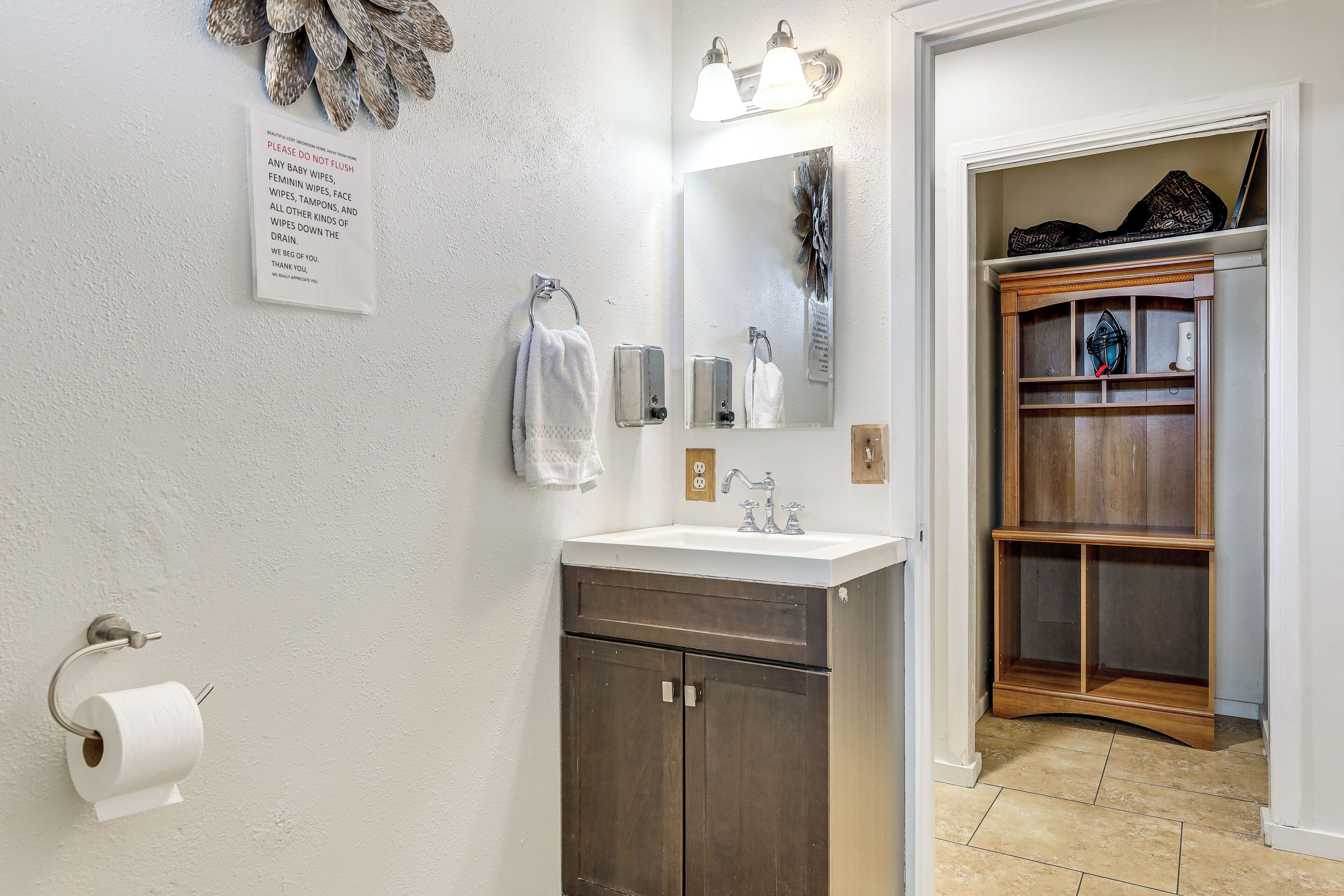 A bathroom with a sink, mirror, towel rack, and a sign instructing not to flush baby wipes.